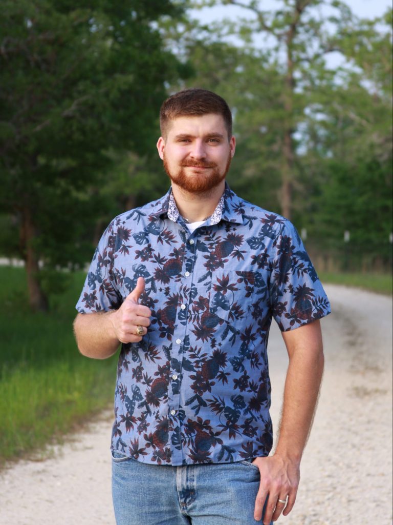 A photo of Josh Tutt giving a thumbs up gesture, wearing a blue floral shirt and standing on a dirt road.