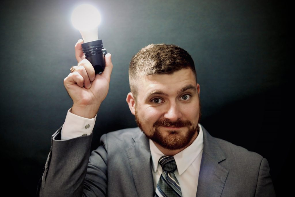 Josh Tutt is standing in front of a dark background, holding an illuminated light bulb above his head with his eyebrows raised. He's having a lightbulb moment.